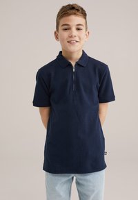 Polo de culoare albastru închis, cu mâneci scurte, având un guler cu fermoar și un material texturat. Asortat cu blugi de culoare albastru deschis.
