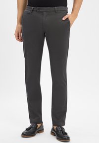 Mężczyzna w ciemnoszarych, teksturowanych spodniach garniturowych z czarnymi mokasynami z frędzlami, z jedną ręką w kieszeni, stojący na jednolitym tle.