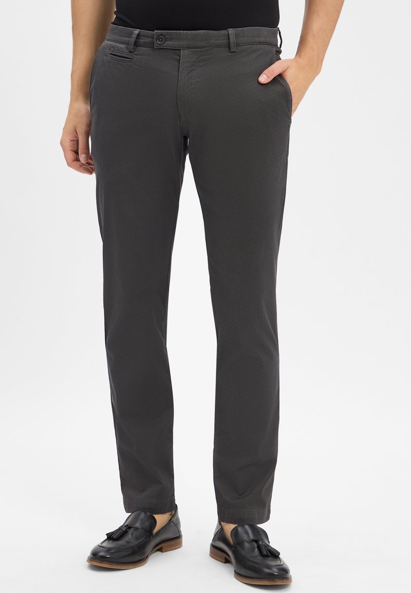 Mężczyzna w ciemnoszarych, teksturowanych spodniach garniturowych z czarnymi mokasynami z frędzlami, z jedną ręką w kieszeni, stojący na jednolitym tle.