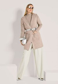 Frau, die eine beige Strickjacke mit silbernem Gürtel, cremefarbene weite Hosen, eine Sonnenbrille, silberne Schuhe trägt und eine kleine silberne Handtasche hält.