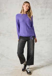 Lila långärmad topp med strukturerad yta, med rund halsringning och justerbar nederkant, i kombination med svarta vida jeans och sneakers.