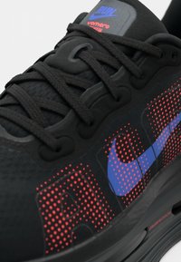 Tenisii negri cu material texturat, având un logo proeminent în culorile albastru și roșu, modele perforate și șirete negre, subliniind un design sportiv.