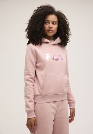 Mujer con cabello rizado que lleva una sudadera con capucha FILA de color rosa claro y pantalones deportivos a juego, mirando hacia un lado sobre un fondo liso.