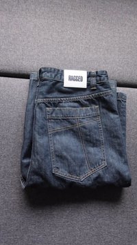 Tamno plave traperice, presavijene s vidljivim šavovima, stražnjim džepom i bijelom etiketom označenom "RAGGED" na pojasu.