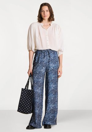 Femme portant un chemisier blanc ample et un pantalon large bleu à motifs, tenant un sac fourre-tout bleu marine avec un petit imprimé blanc, debout sur un fond uni.