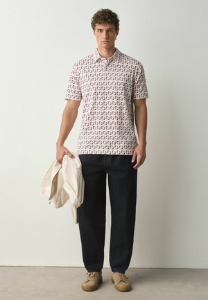 Jeune homme debout, portant une chemise à manches courtes à motifs, un pantalon noir, des chaussures beiges et tenant une veste beige pliée dans une main.