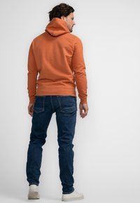Felpa arancione con tasca a marsupio, polsini aderenti e cappuccio con coulisse, abbinata a jeans blu scuri e sneakers bianche.
