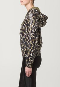 Hupparillinen takki kiiltävällä pinnalla, jossa monivärinen abstrakti kuvio vihreän, ruskean ja mustan sävyissä. Kuminauhakuja helmassa ja sivutaskut.