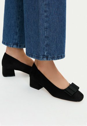 Piedi con scarpe basse in camoscio nero con tacco a blocco e fiocchi, abbinate a jeans blu di denim larghi e cropped.