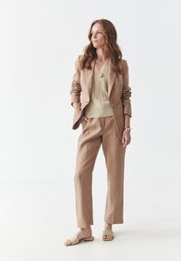 Costume en lin brun clair avec coupe décontractée, pantalon assorti, haut texturé beige et sandales à glissière tan. Un collier pendentif en or accentue le look.