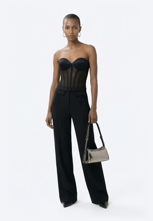 Γυναίκα που φορεί ένα μαύρο διάφανο κορσέ χωρίς τιράντες και μαύρο παντελόνι με φαρδιά πόδια, κρατώντας μια λαμπερή ασημένια τσάντα με αλυσίδα.