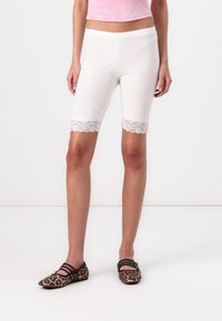 Hvide strækshorts med blondekant i kanten, tætsiddende design, glat tekstur. Båret med mønstrede flade sko.