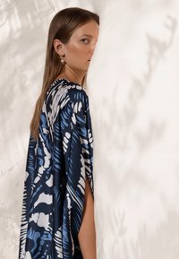 Tissu bleu marine avec des motifs abstraits blancs et bleu clair qui tombent élégamment. Modèle partiellement caché par un fond blanc, avec des manches longues.
