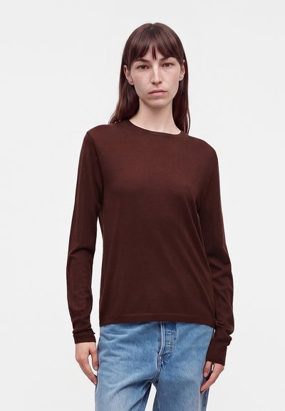 Femme aux cheveux bruns raides portant un t-shirt marron uni à manches longues et un jean bleu clair, debout devant un fond blanc.