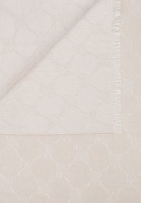 Textil color crema con un sutil patrón floral geométrico, una esquina doblada para revelar la textura de la tela y el detalle del borde de flecos.