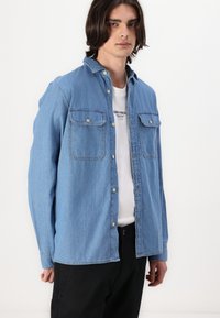Lichtblauwe denim shirt met twee borstzakken, knoopsluiting aan de voorkant, lange mouwen en een rechte onderkant. Draag dit over een wit t-shirt.