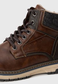 Braune Leder-Stiefelette mit grauen Stoffakzenten, runden Ösen, dunklen Schnürsenkeln und einer strukturierten, robusten Sohle. Markenlogo sichtbar.