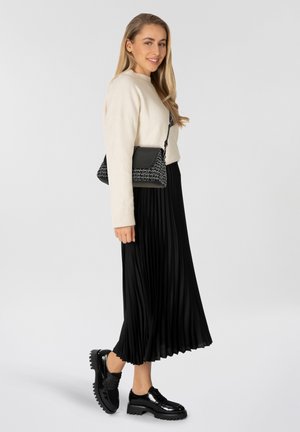 Cremfarbener Pullover, schwarzer plissierter Midirock, glänzende schwarze Schuhe mit dicken Sohlen und eine schwarz-weiße geflochtene Tasche mit Tragegurt.