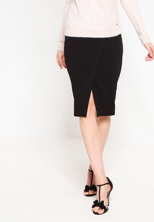 Jupe crayon noire avec fente latérale. Le tissu est lisse avec un design ajusté. Portée avec des escarpins à lanière avec un détail en nœud.