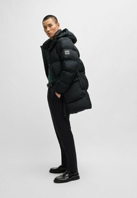 Black puffer jacket with a hood, features a soft texture, quilted design, and a logo patch. Worn over a green top and paired with dark pants.