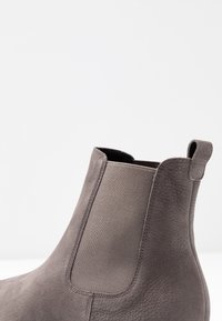 Šedá semišová kotníková bota s elastickou boční částí a poutkem, s texturovaným povrchem a minimalistickým designem. Klasický styl Chelsea bot.
