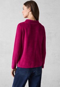 Maglietta fucsia a maniche lunghe in morbido tessuto a costine, con colletto rotondo e spacchi laterali, indossata con jeans blu scuro. Mostrata vista da dietro.