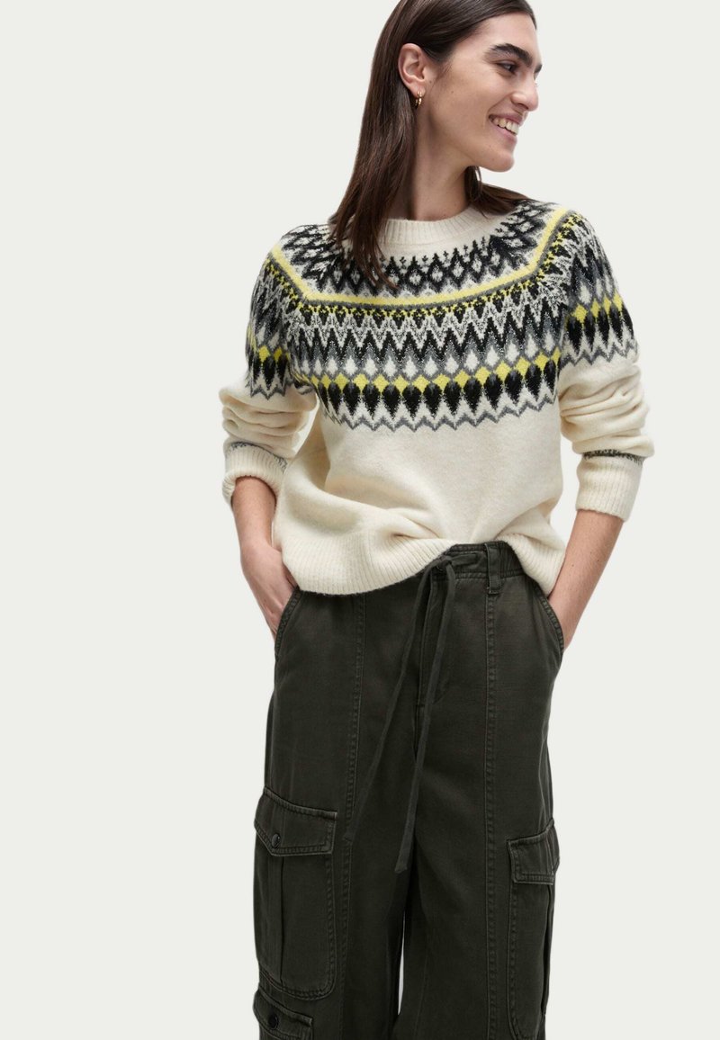 Krämfärgad stickad tröja med geometriska mönster i svart, gult och grått, kombinerad med mörkgröna cargobyxor med sidofickor.