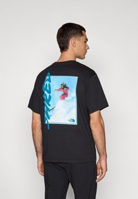 Black cotton t-shirt featuring a colorful skier graphic on the back, with bold turquoise text and The North Face logo in the corner.