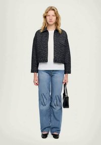 Svart mönstrad kort jacka över en vit skjorta, tillsammans med blå jeans med logodesign och en svart handväska. Släta texter och avslappnad passform.