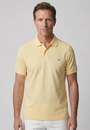 Mann trägt ein kurzärmeliges hellgelbes Poloshirt mit kleinem Diamant-Logo auf der Brust, steht vor einem schlichten Hintergrund.