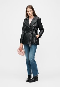 Frau in schwarzer Lederjacke mit Gürtel, blauen Jeans mit geradem Bein und schwarzen Knöchelstiefeln, hält eine kleine pinke Clutch-Tasche.