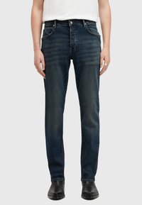 Jeans de mezclilla azul oscuro con un ajuste slim, que cuentan con bolsillos frontales, costuras visibles y un cierre de botón. Llevados con botas negras hasta los tobillos.