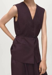 Bluzka bez rękawów o głębokim dekolcie w kształcie litery V, wykonana z ciemnofioletowej tkaniny, z wcięciem w talii i dwoma bocznymi kieszeniami o gładkim wykończeniu.