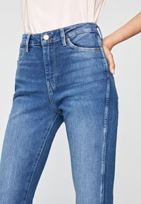 Högmidjade blå denimjeans med klassisk passform, med en framficka och synliga sömdetaljer, slät textur och lätt blekning.