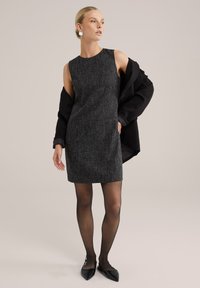 Melna bezpiedurkņu kleita ar teksturētu audumu, kas satur divas priekšējās kabatas. Modelis valkā caurredzamas melnas zeķbikses un melnas norādītās kurpes.