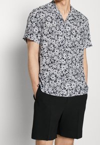 Hombre vistiendo una camisa de botones de manga corta en blanco y negro con estampado floral, combinado con pantalones cortos negros lisos hasta la rodilla, con las manos en los bolsillos.