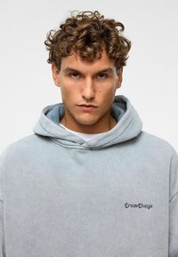 Lichtgrijze hoodie gemaakt van zachte stof, met een voorzak en geborduurd "Create Change" logo op de borst.