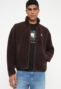 Jachetă din fleece maro cu guler înalt, închidere cu fermoar și buzunare laterale, având un material texturat, moale și o margine contrastantă.