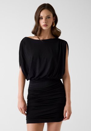 Jonge vrouw in een zwarte mini-jurk met plooitjes, een losse boothals en gouden oorringen, staand tegen een effen lichte achtergrond.