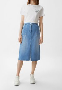 Kobieta w białej koszulce z sercem z napisem "miłość", w średniej długości niebieskiej dżinsowej spódnicy z przednimi kieszeniami oraz w białych sneakersach.