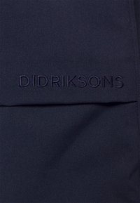 Tmavě modrá tkanina s vyšitým nápisem "DIDRIKSONS" v odpovídající nitě nad vodorovným švem.