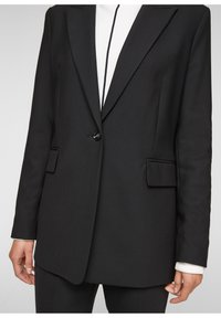 Svart skräddarsydd blazer med enstaka knappstängning, slagkrage, framfickor och slät textur. Bärs över en vit topp.