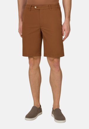 Mand iført brune skræddersyede shorts og grå slip-on sko, stående med afslappede arme langs siden mod en ensfarvet baggrund.