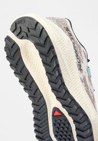 Σόλα παπουτσιού για ορεινό τρέξιμο με βαθείς μαύρους αυλακωτούς, μπεζ μεσαία σόλα και κόκκινη τριγωνική λεπτομέρεια, σχεδιασμένη για πρόσφυση και ανθεκτικότητα.