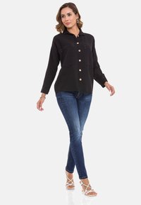 Zwarte blouse met lange mouwen en knopen, voorzien van twee voorzakken, houten knopen, gedragen met blauwe skinny jeans en witte hakken.