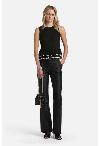 Femme portant un haut noir sans manches à volants avec bordure blanche, pantalon noir à jambes larges, talons ouverts noirs, tenant un sac à main noir avec chaîne dorée.