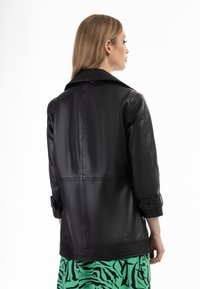 Schwarze Lederjacke mit Kragen und umgeschlagenen Ärmeln. Glatte Textur und taillierter Schnitt, kombiniert mit einem Rock mit grün-schwarzem Muster.