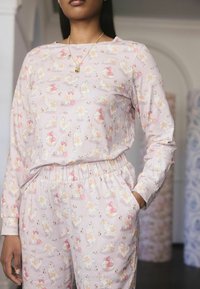 Een vrouw modelleert pastelkleurige pyjama's met speelse kattenontwerpen, met één hand in haar zak en een delicaat kettingtje om haar nek.