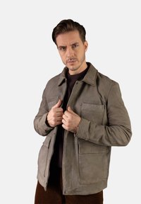 Šedá kožená bunda s límcem, čtyřmi p�ředními kapsami a zipovým zapínáním. Hladká textura s matným povrchem, nošena přes tmavý svetr.