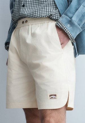 Persona con pantalones cortos color crema con aberturas laterales y un parche con el logo de Namushka, combinado con una camisa estampada y una chaqueta azul claro.